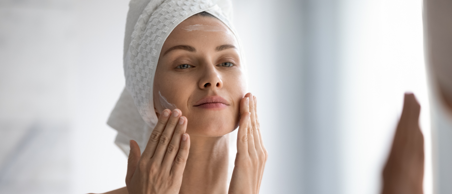 Žena v koupelně před zrcadlem si aplikuje pleťový krém.